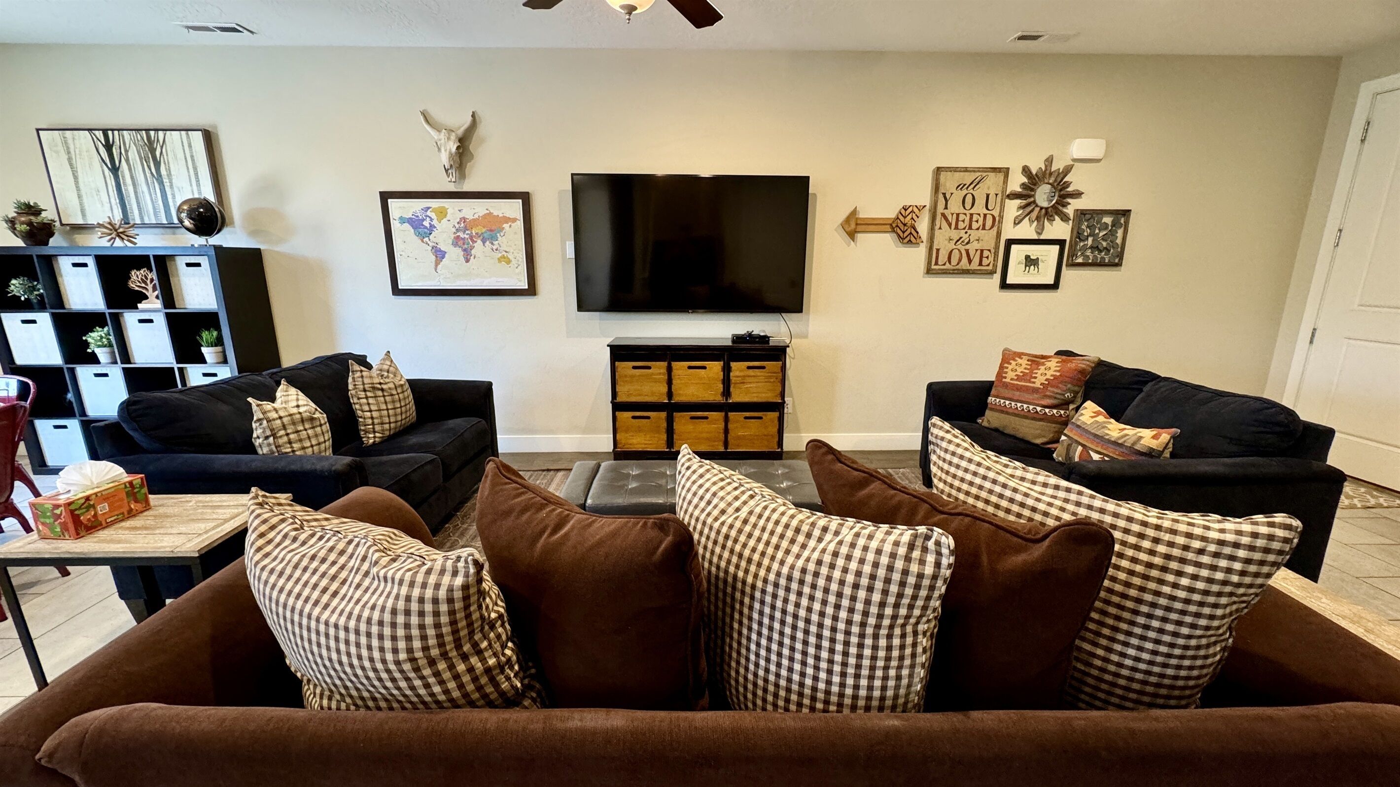 A living room with two black sofas, a brown couch, a flat-screen TV, a world map, a cow skull, and various wall decorations.