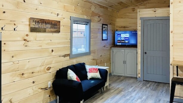 A cozy cabin interior with a wooden ceiling, a black sofa, a television, and a door. - UTV/ATV PARADISE! A/C and heat (Huntsville)