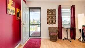 Una habitación con una pared roja, una puerta que da al exterior, una silla de mimbre, una mesita con una lámpara y un ventanal con cortinas.