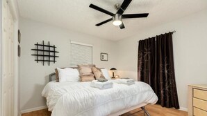Un dormitorio con cama, ventilador de techo, mesita de noche, cómoda y ventana con persianas.