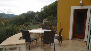Un patio al aire libre con una mesa redonda y cuatro sillas, un banco de piedra y una pared amarilla con una puerta.