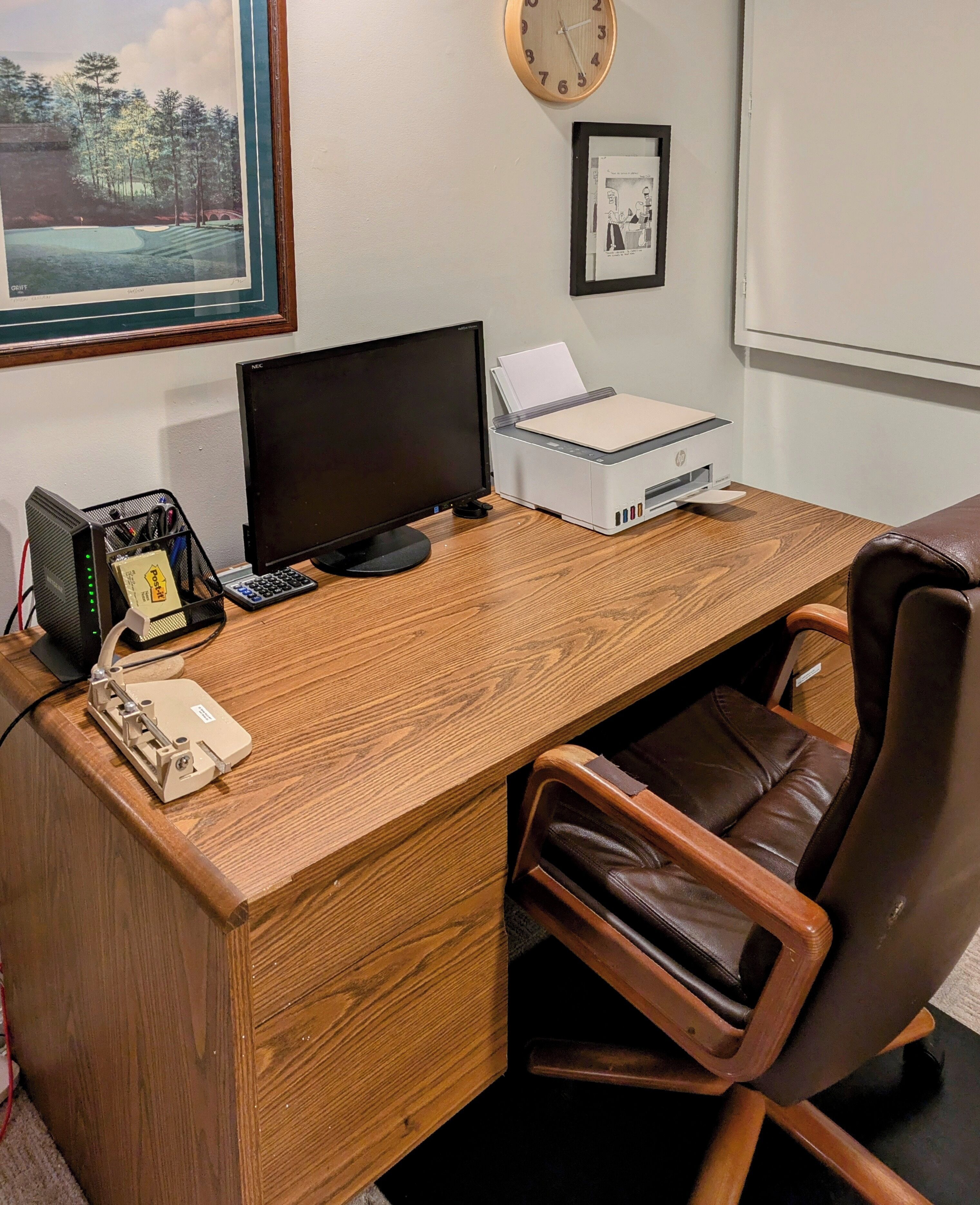 Un escritorio de madera con una computadora, una impresora y un teléfono. Una silla está colocada frente al escritorio.