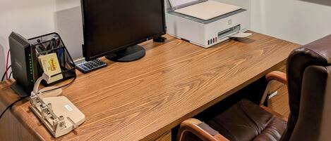Un escritorio de madera con una computadora, una impresora y un teléfono. Una silla está colocada frente al escritorio.