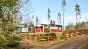 Een klein, rood houten huisje met een glazen tuinkamer, omgeven door een houten hek en een dicht bos.