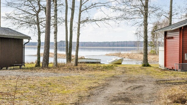 Een grindpad leidt naar een meer met boten, omringd door bomen en twee gebouwen.