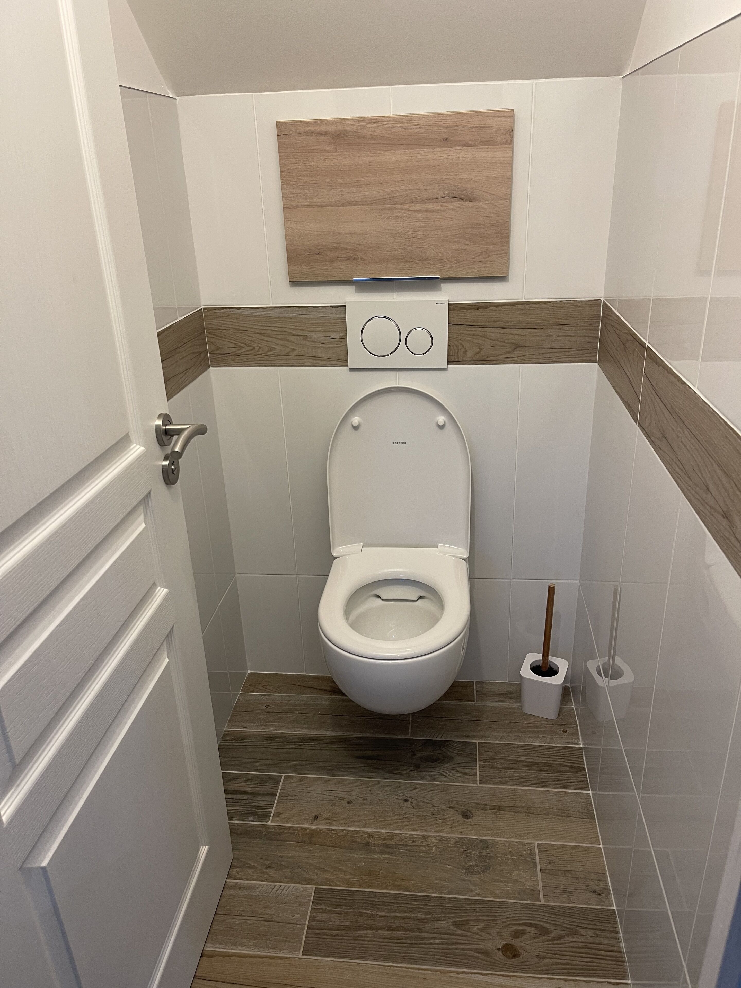 Un baño con inodoro, un panel de madera sobre él y un botón de descarga en la pared.