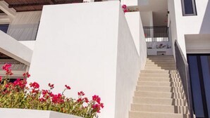 A modern multi-story building with white exterior walls, a staircase leading to the upper floors, and pink flowers in the foreground. - Oceanfront at Cerritos Beach • Infinity Pool+Spa (Los Cerritos)