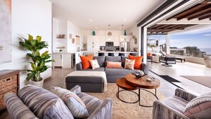 A modern living room with a grey sofa, ottoman, and coffee table, a kitchen in the background, and a balcony with outdoor furniture. - Oceanfront at Cerritos Beach • Infinity Pool+Spa (Los Cerritos)