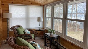 Una habitación acogedora con paredes y techo de madera, un sillón de mimbre con cojines, una mesita con una lámpara y un ventanal con vistas a árboles.