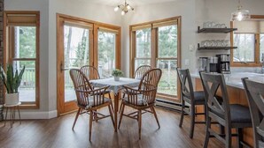 Comedor con una mesa redonda y cuatro sillas, ventanas amplias y una zona de cocina con estantes y una lámpara colgante.