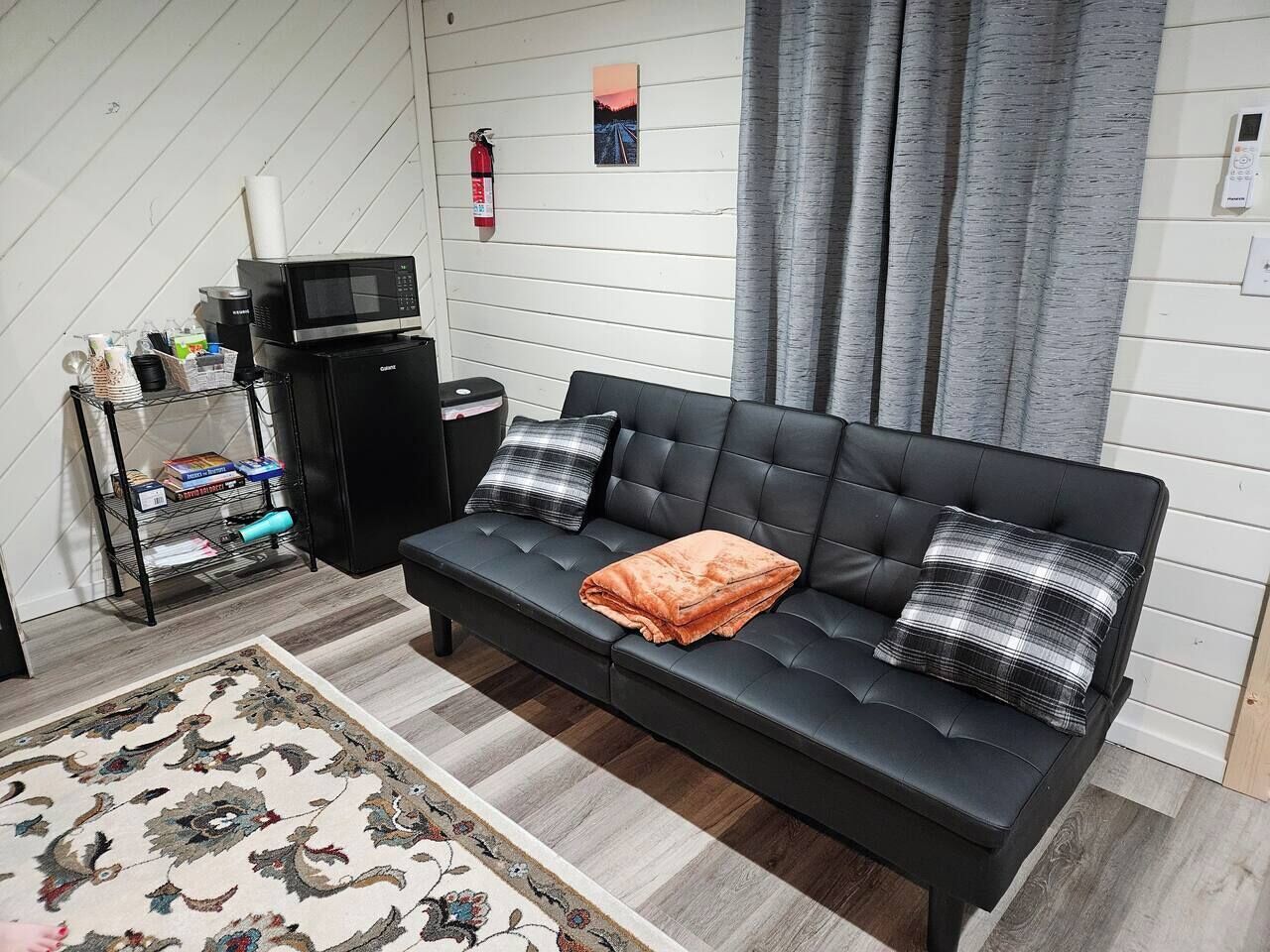 Un salon moderne avec un canapé en cuir noir, deux coussins, une petite table basse, un four à micro-ondes, un extincteur et une étagère contenant divers objets.