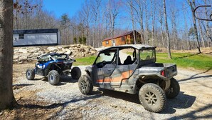 Two off-road vehicles parked on a gravel path with a wooden cabin and a metal structure in the background. - UTV/ATV PARADISE! A/C and heat  (Huntsville)