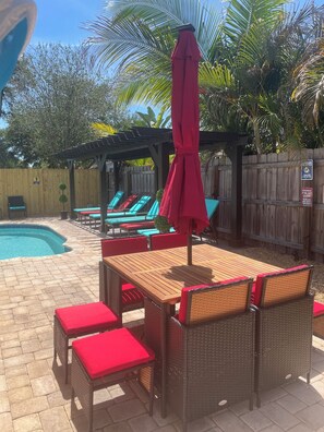 A poolside area with a wooden table and wicker chairs, a red umbrella, and lounge chairs. - HOME SWEET HOME!☀️ HUGE HOT TUB. HEATED POOL! 7 miles to beach 4BD 2BA updated! (Seminole)