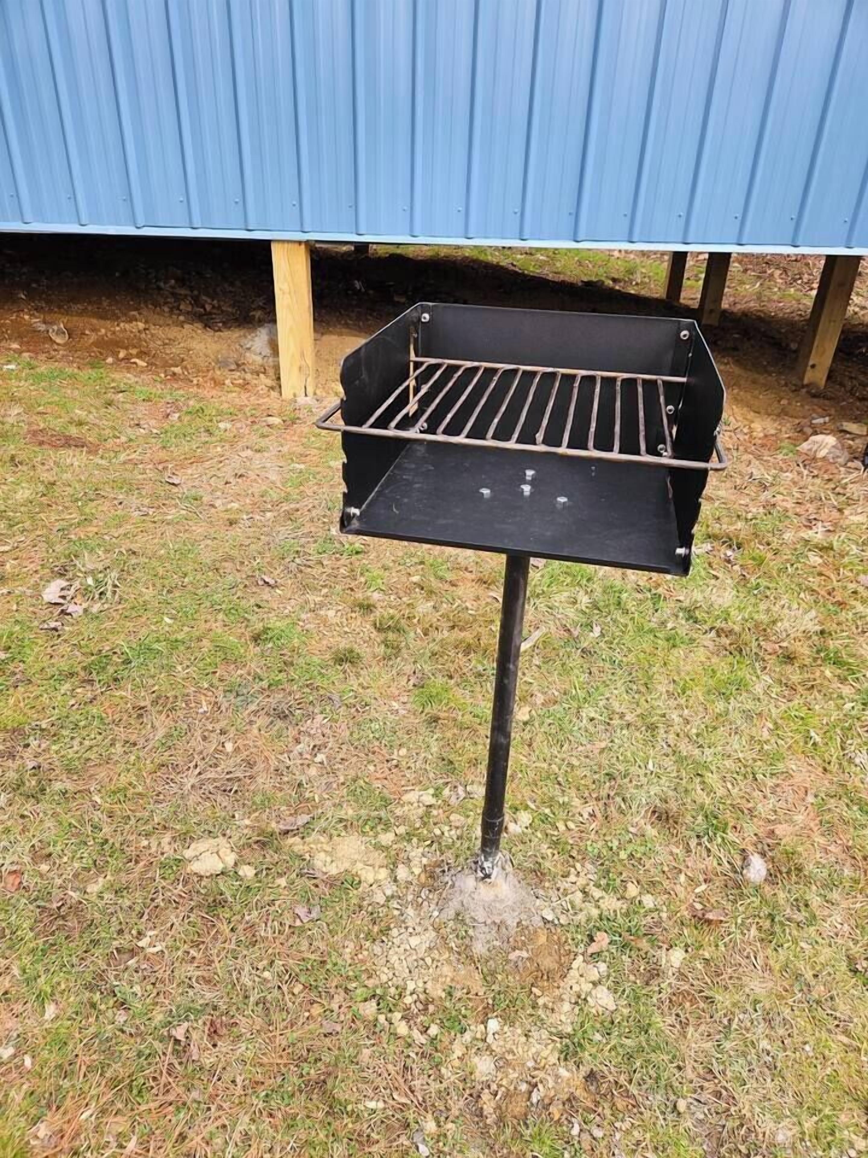 Een draagbare barbecue met metalen rooster, gemonteerd op een potenstandaard, buiten geplaatst op een gazon met een blauwe metalen constructie op de achtergrond.