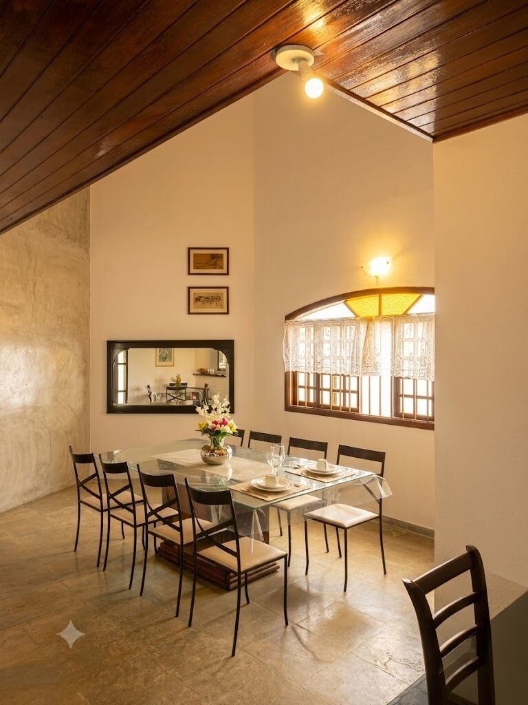 Un comedor con una mesa de vidrio, sillas, un jarrón de flores, un espejo y una ventana con cortinas.