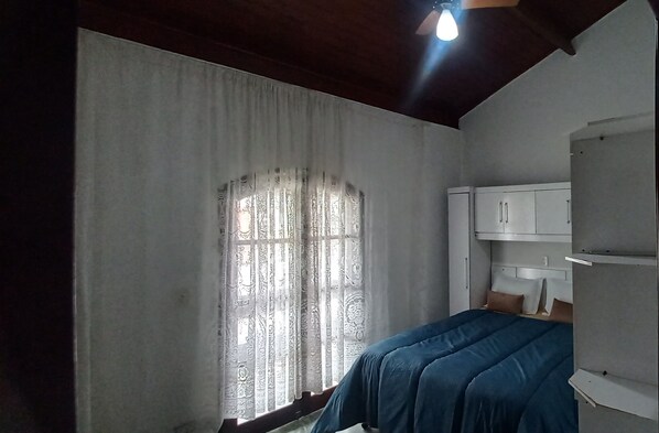Una camera da letto con un letto singolo, una finestra con tende a motivi, una ventola a soffitto e un armadio bianco.