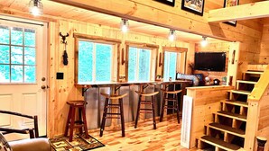 Interior de uma cabana de madeira com espaço de bar, escadas de madeira e uma televisão.