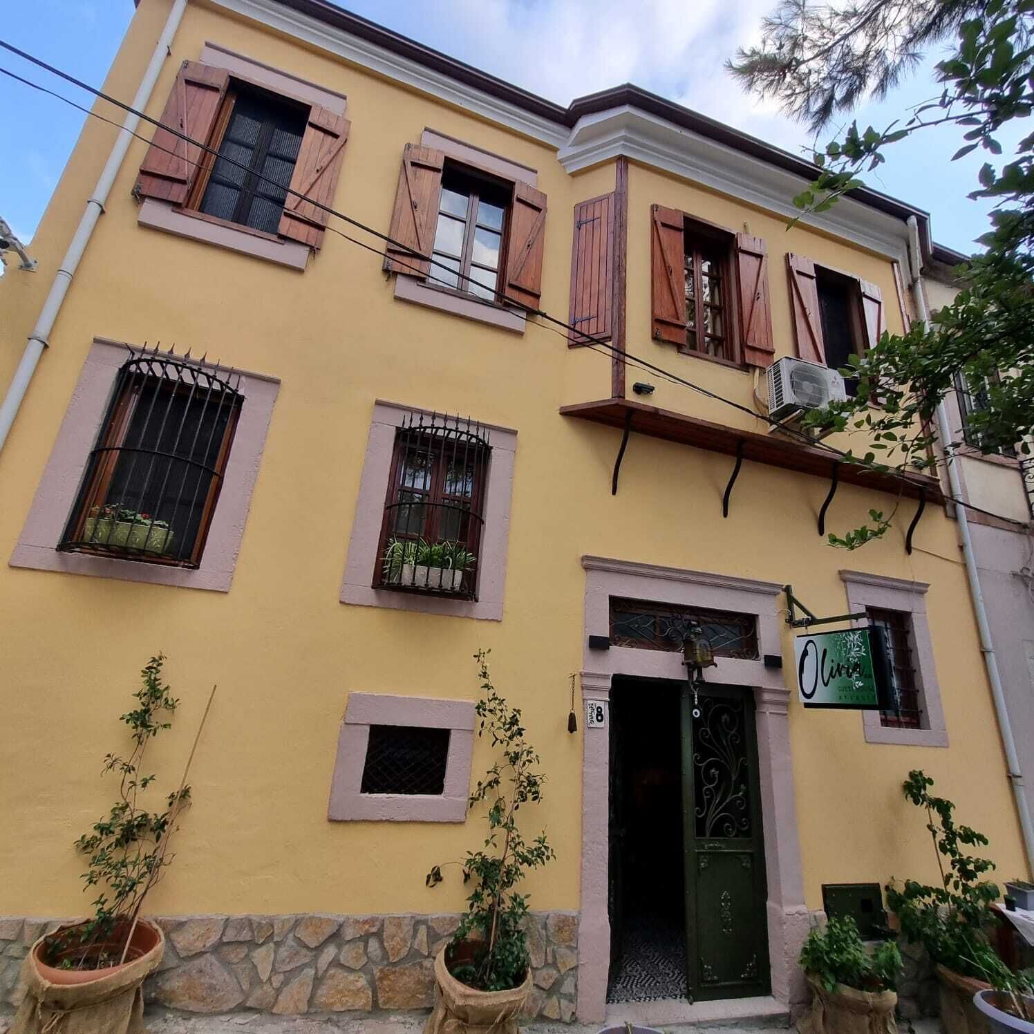 Un edificio amarillo con múltiples ventanas, algunas con contraventanas, y una puerta con un enrejado de hierro decorativo. Hay plantas en macetas en la base del edificio.