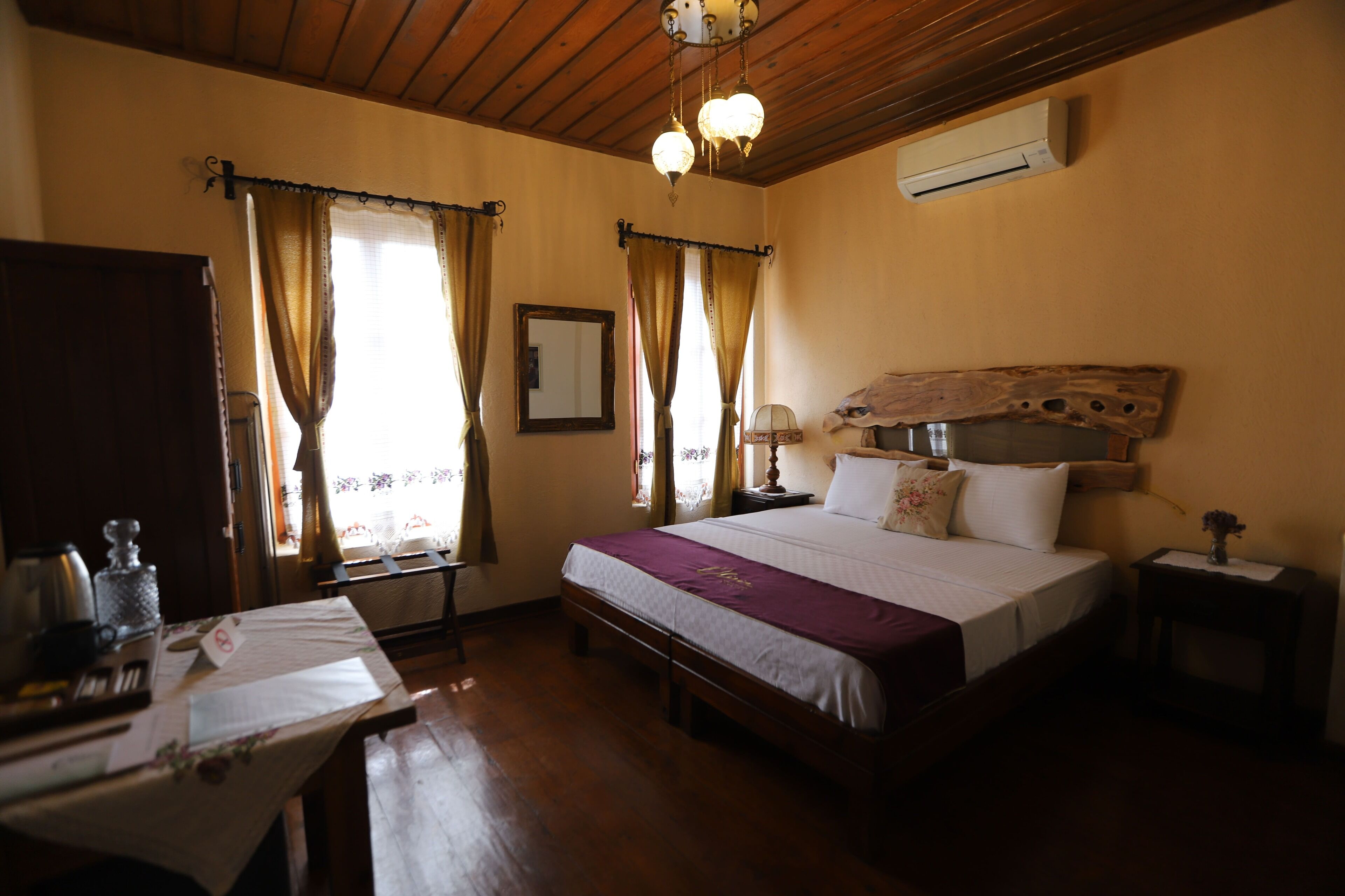 A bedroom with a wooden bed, a bedside table, a nightstand, a mirror, and a window with curtains.
