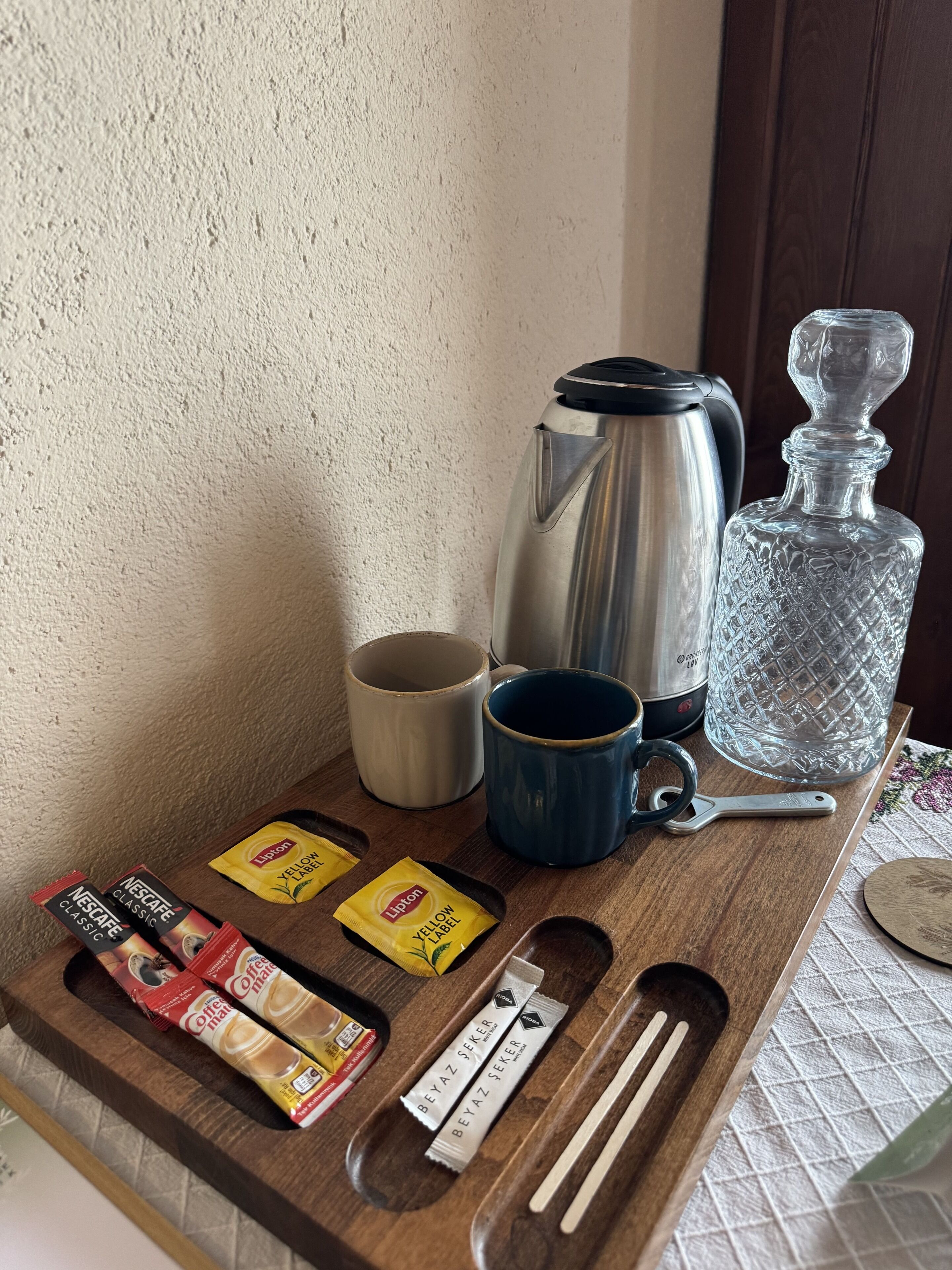 Een houten dienblad met koffiebereiding accessoires, waaronder een Nescafe Classic koffiecup, Lipton Yellow Label theezakjes, Beyaz Seeker koffiecups, een blauwe mok, een roestvrijstalen koffiepot en een kristallen karaf.