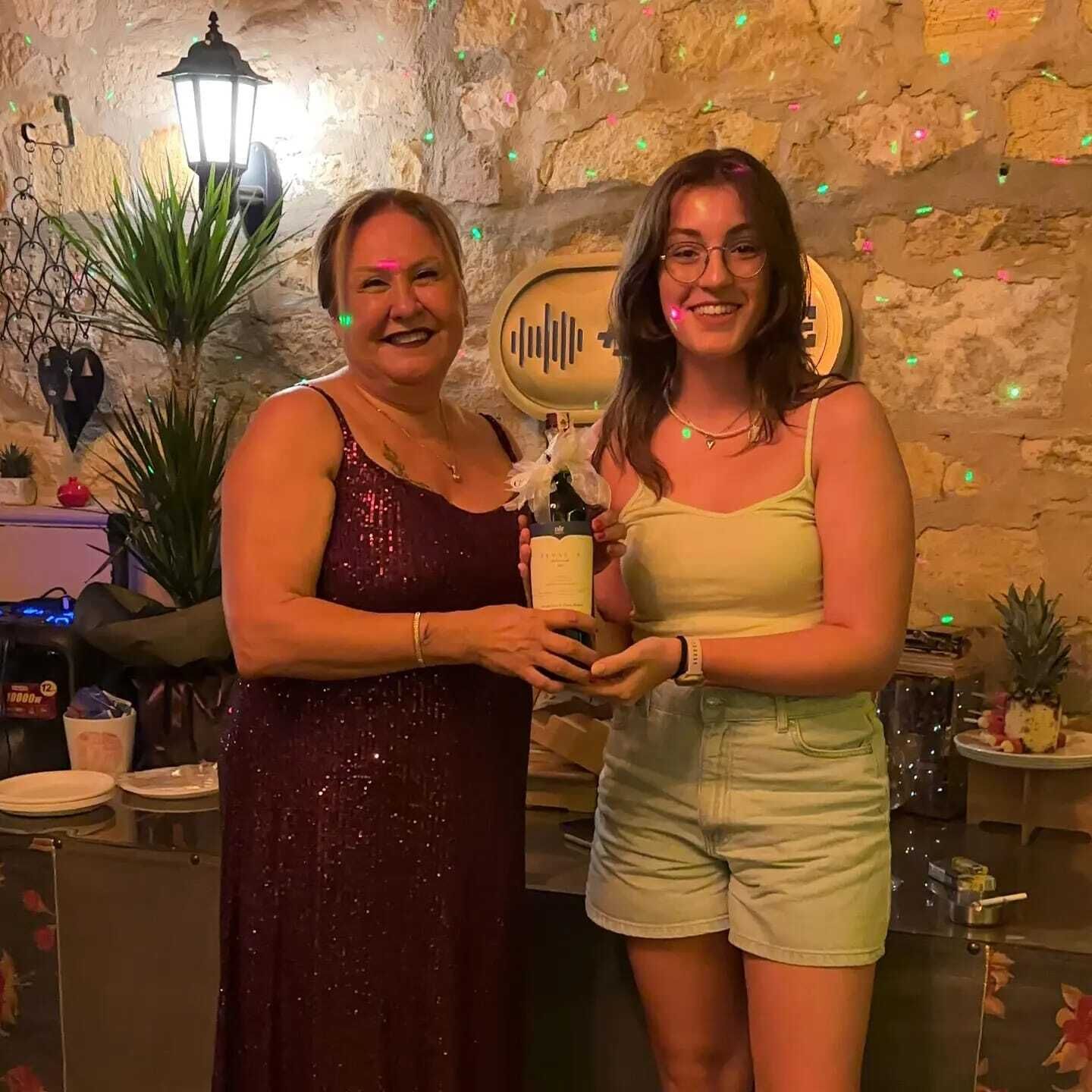 Two people standing in a room with stone walls, a table with plates and a bottle, and a decorative plant.