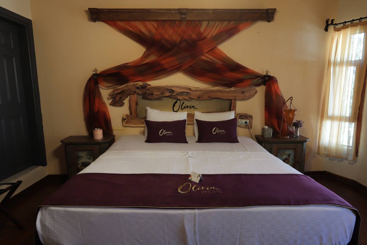 Een bed met hoofdbord versierd met een rustiek ontwerp, twee kussens met de naam 'Olivia' en een sprei met dezelfde naam.
