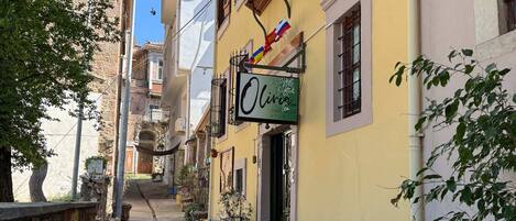 A narrow street with buildings on either side, a sign for 'Olive Guest House', outdoor seating with tables and chairs, and potted plants.