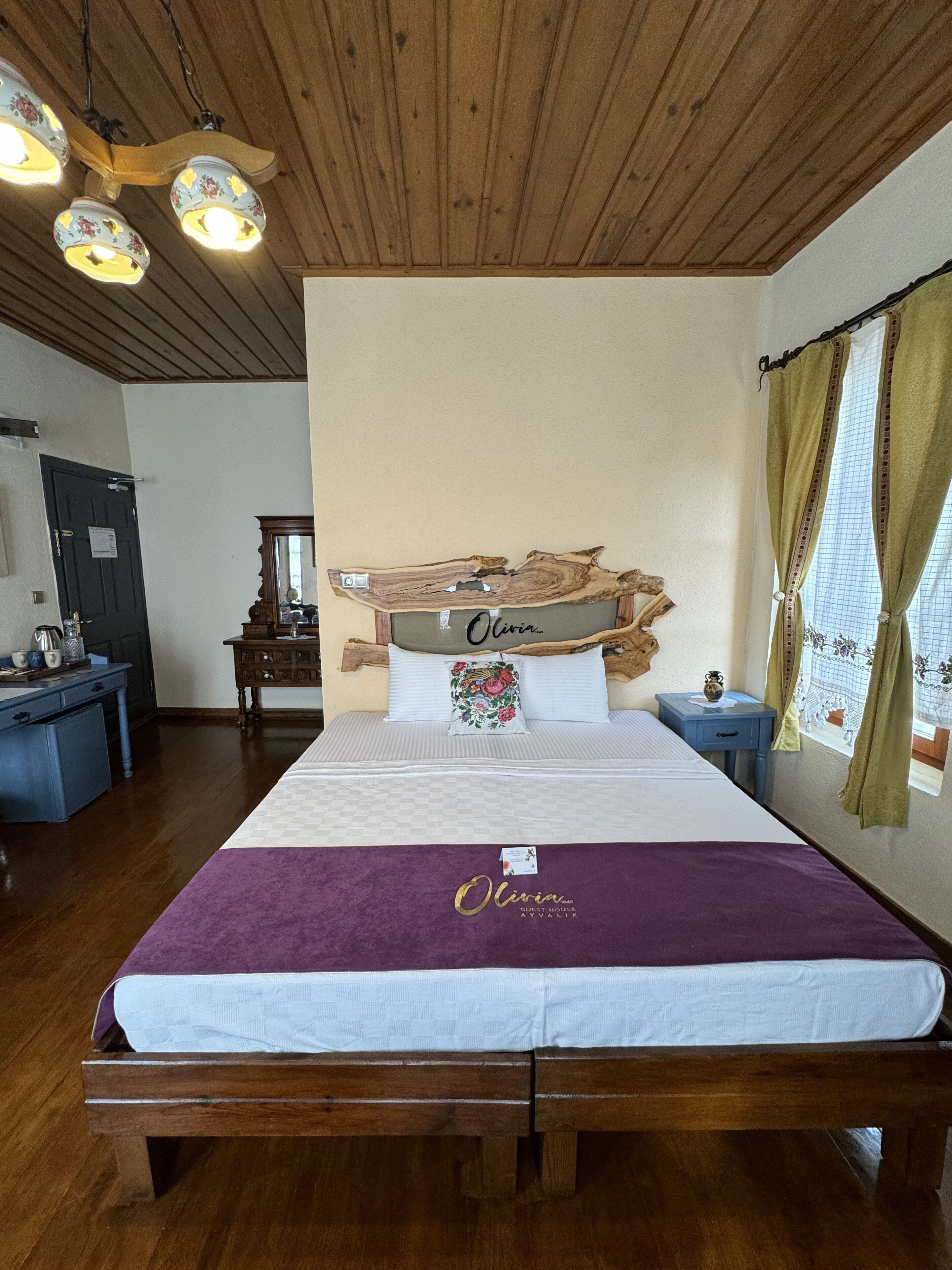 Une chambre d’hôtes avec un plafond en bois, un lit recouvert d’une courtepointe violette, une table de chevet, un miroir et une fenêtre avec des rideaux.