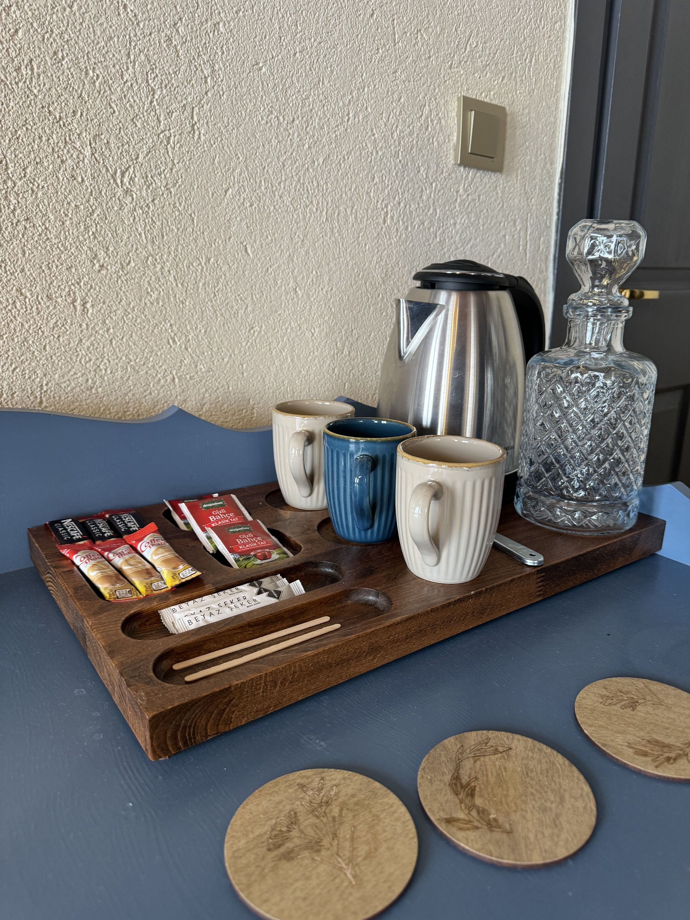 Una bandeja de madera con tazas de café, una cafetera, un decantador de cristal y aperitivos envasados sobre una mesa azul junto a una pared con textura.