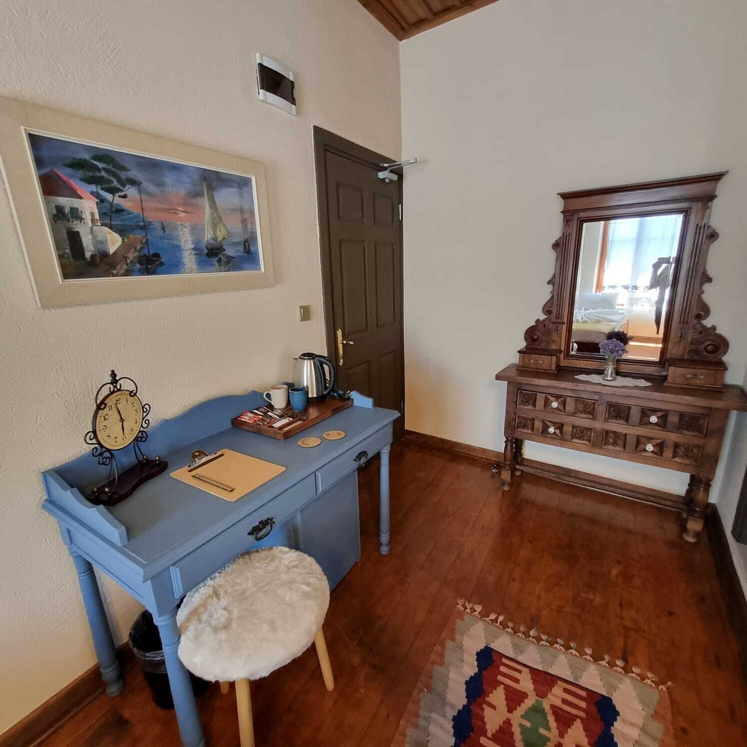 A room with a blue desk, a white fluffy stool, a wooden dresser, a mirror, a painting, and a patterned rug.