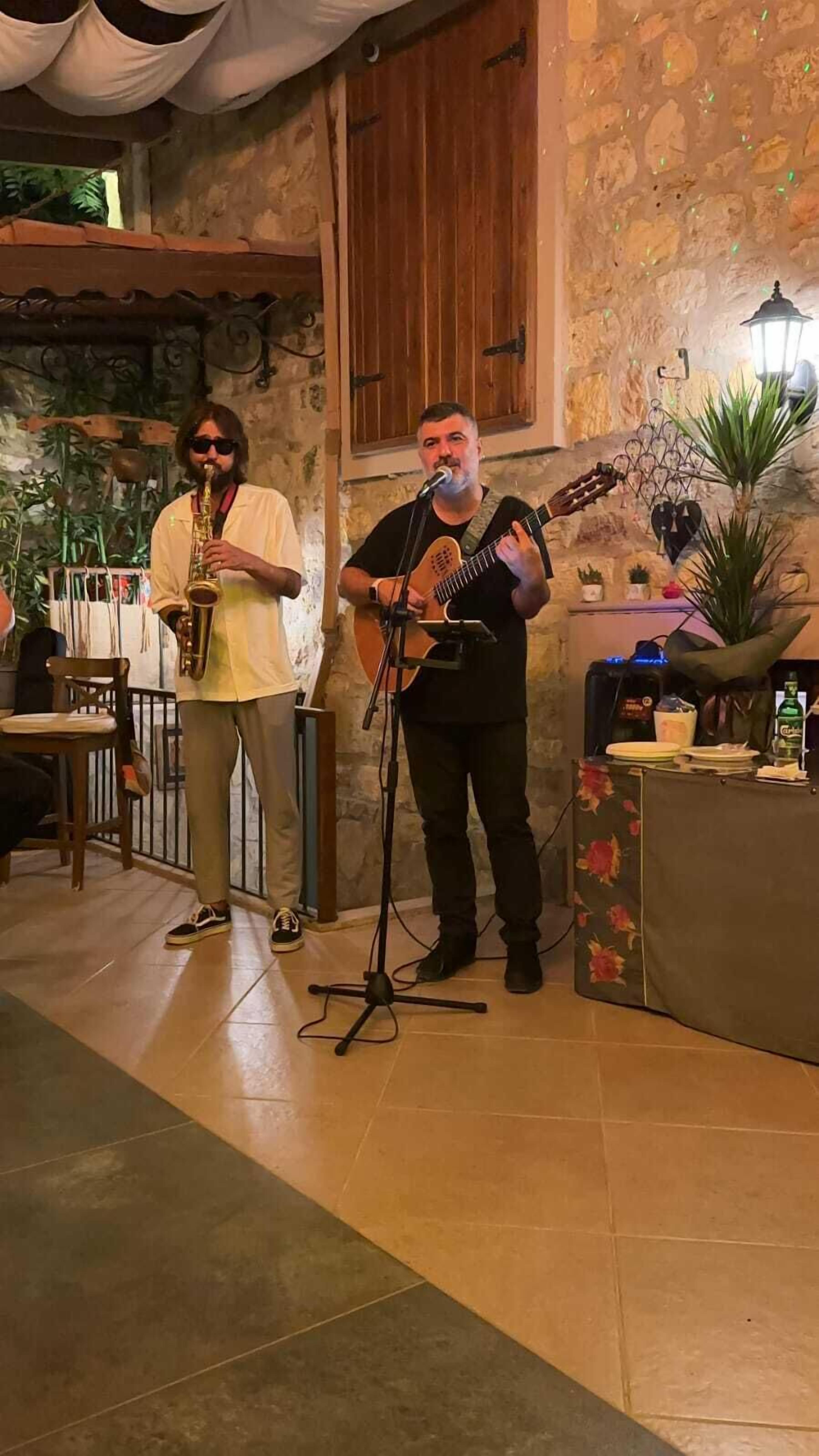 A musician playing a guitar and a saxophone in a rustic room with stone walls and a wooden door.