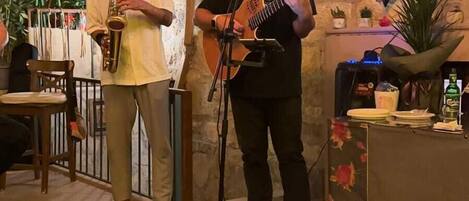 A musician playing a guitar and a saxophone in a rustic room with stone walls and a wooden door.