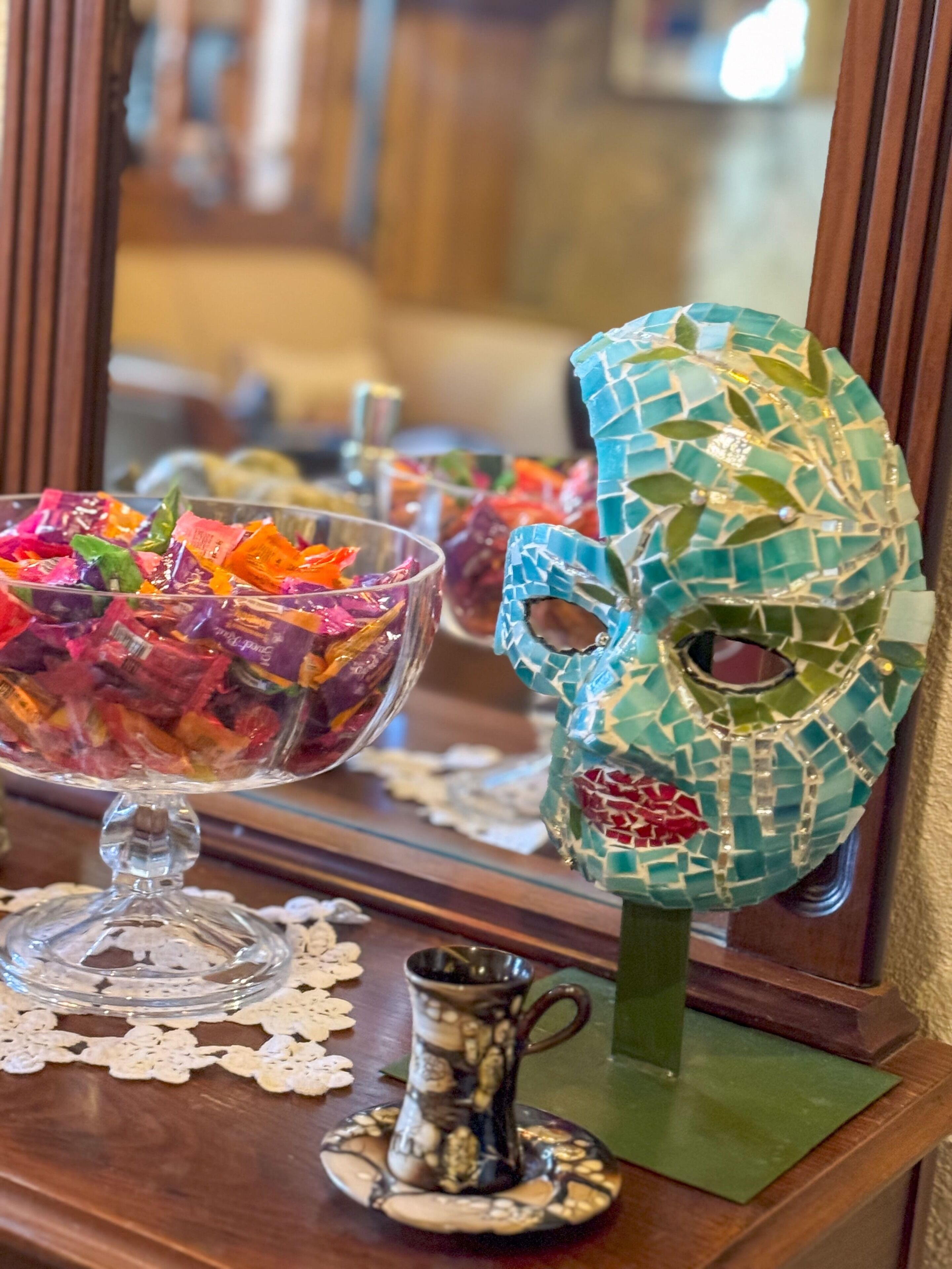 Una ciotola di vetro piena di caramelle colorate su un tavolo di legno, con una maschera decorativa e una piccola brocca.