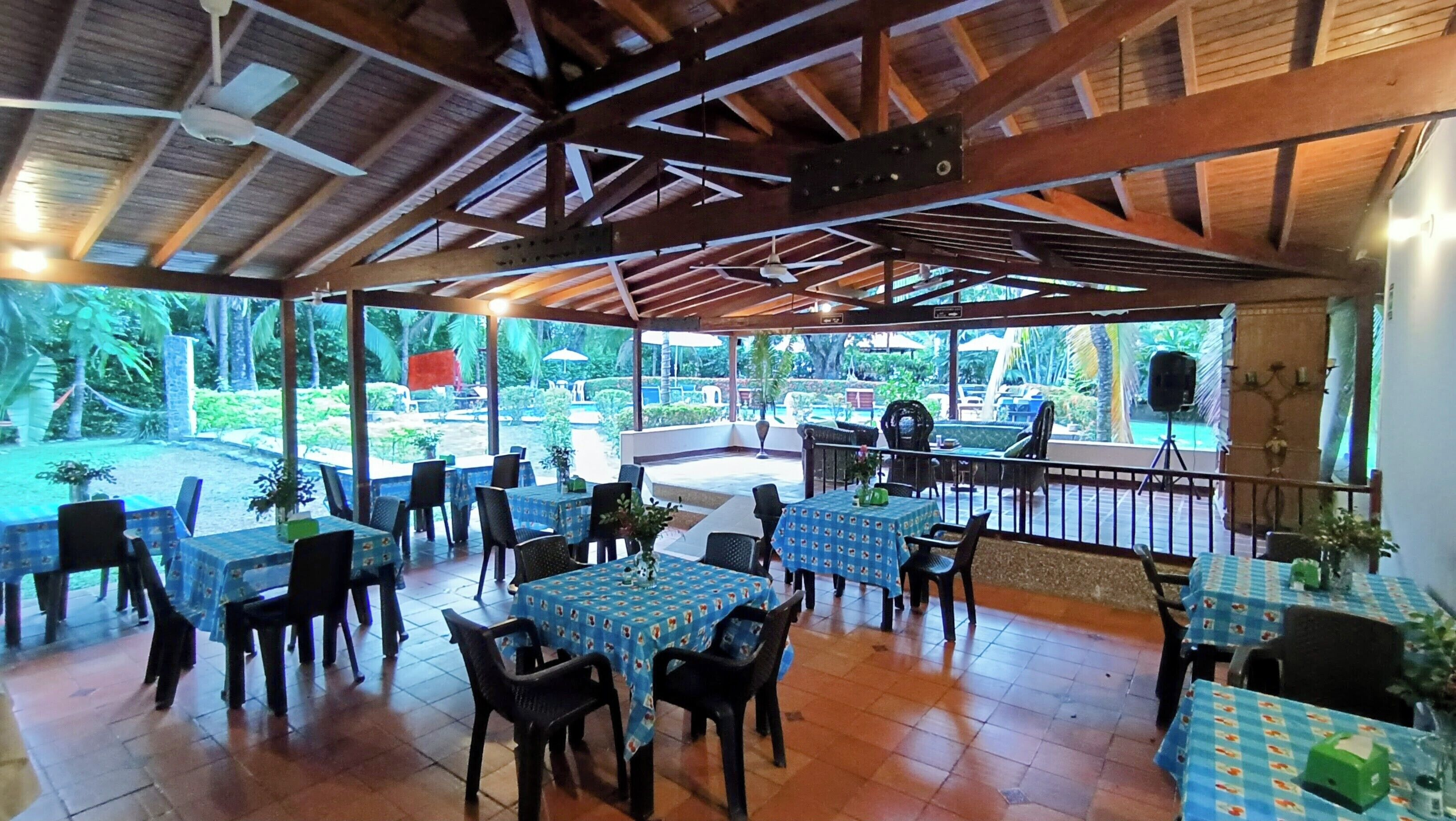 Un espace de restauration en plein air, avec des tables et des chaises, un toit abrité et une vue sur une piscine et de la verdure.