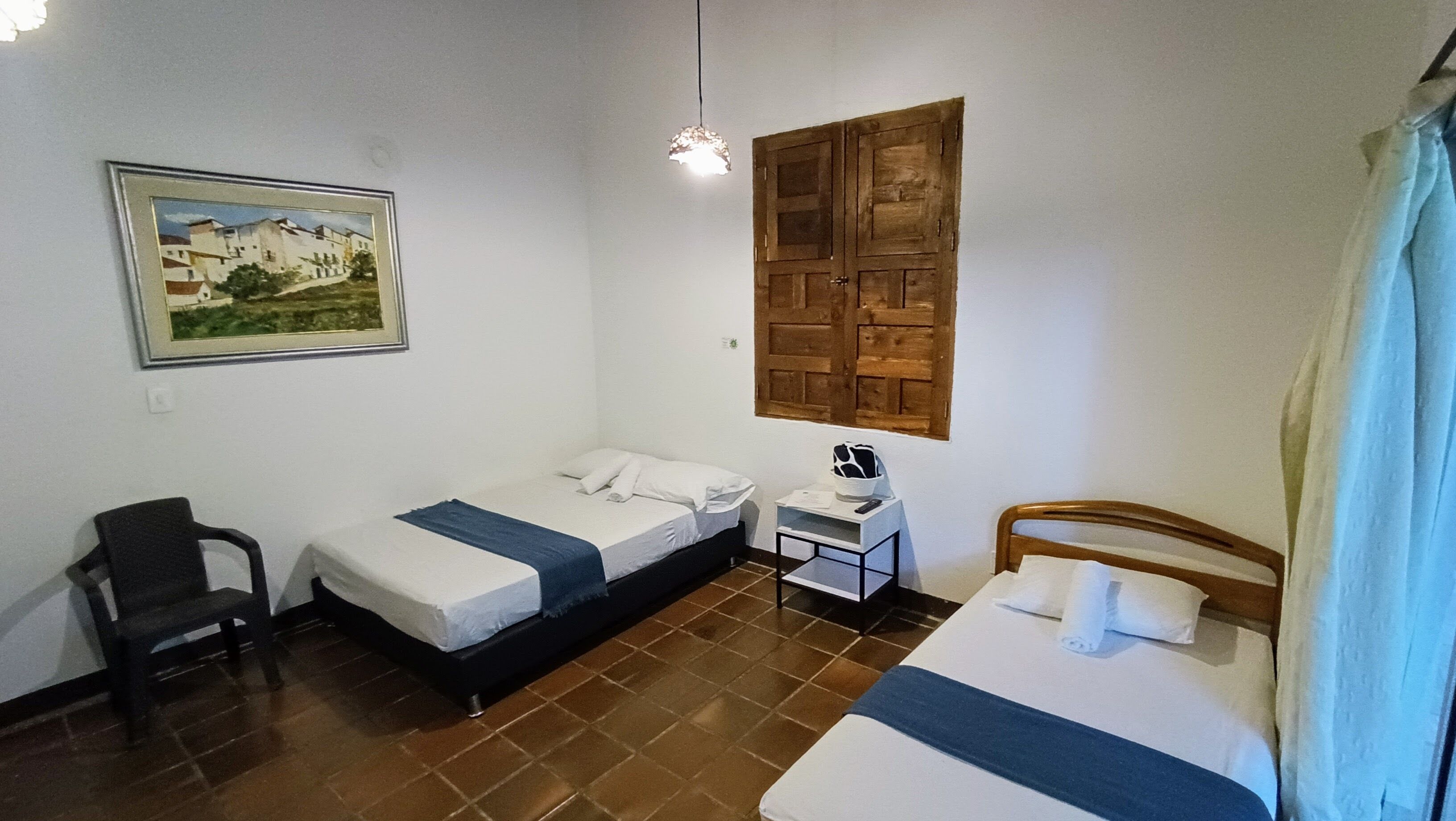 A small, clean room with two beds, a chair, a nightstand, and a painting on the wall.