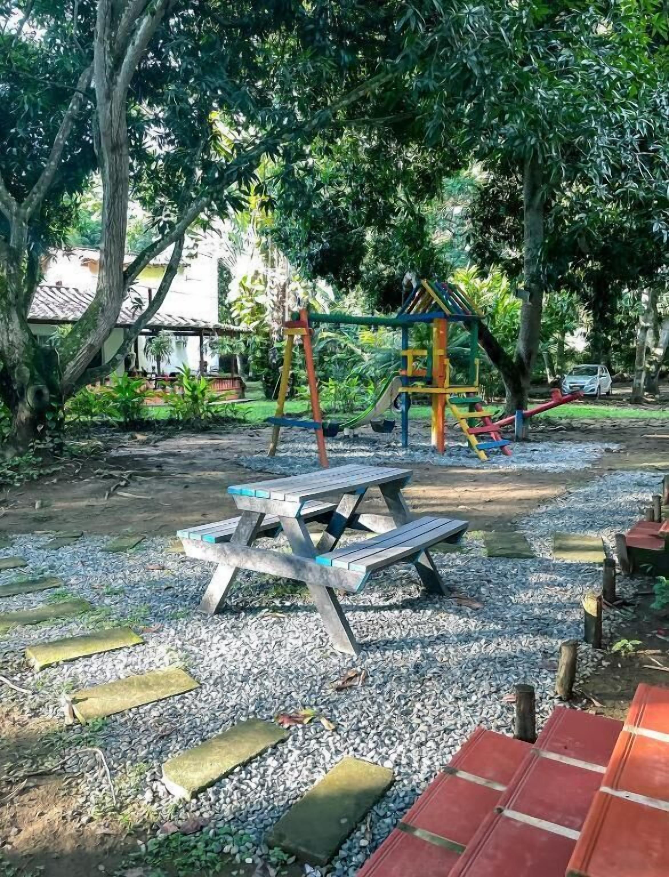 Un parque infantil con tobogán y columpios, rodeado de árboles y grava.