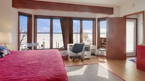 Quarto com uma cama grande, escrivaninha e cadeira. Janelas amplas oferecem vista para a praia.