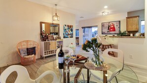 A modern living room with a dining area, featuring a glass table, white chairs, a wine bottle, and a vase with flowers.