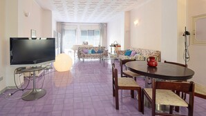 Un salon doté d’un téléviseur à écran plat, d’une table de salle à manger ronde avec des chaises, de canapés à motifs et d’un grand luminaire.