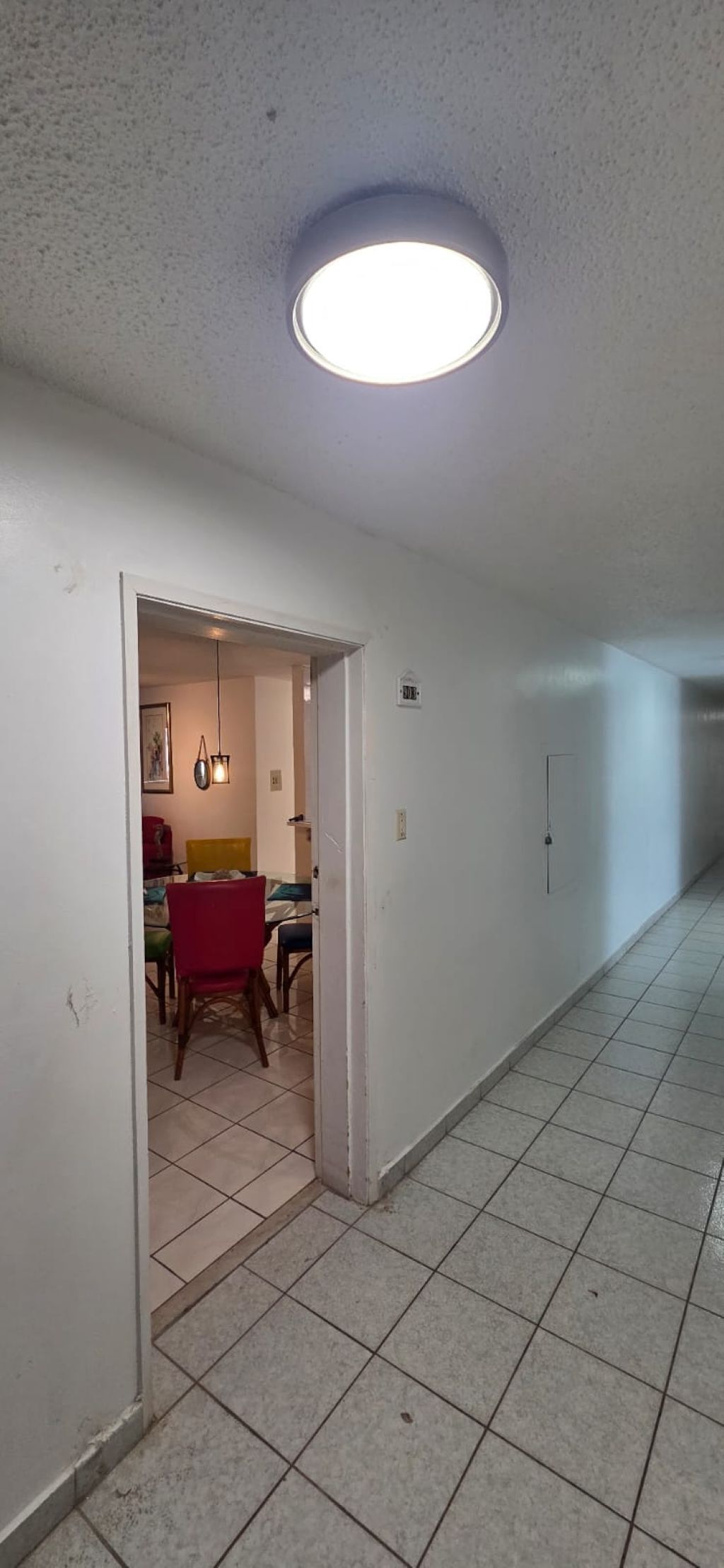 Un pasillo con piso de baldosas, una puerta que da a un comedor con mesa y sillas, y una lámpara en el techo.