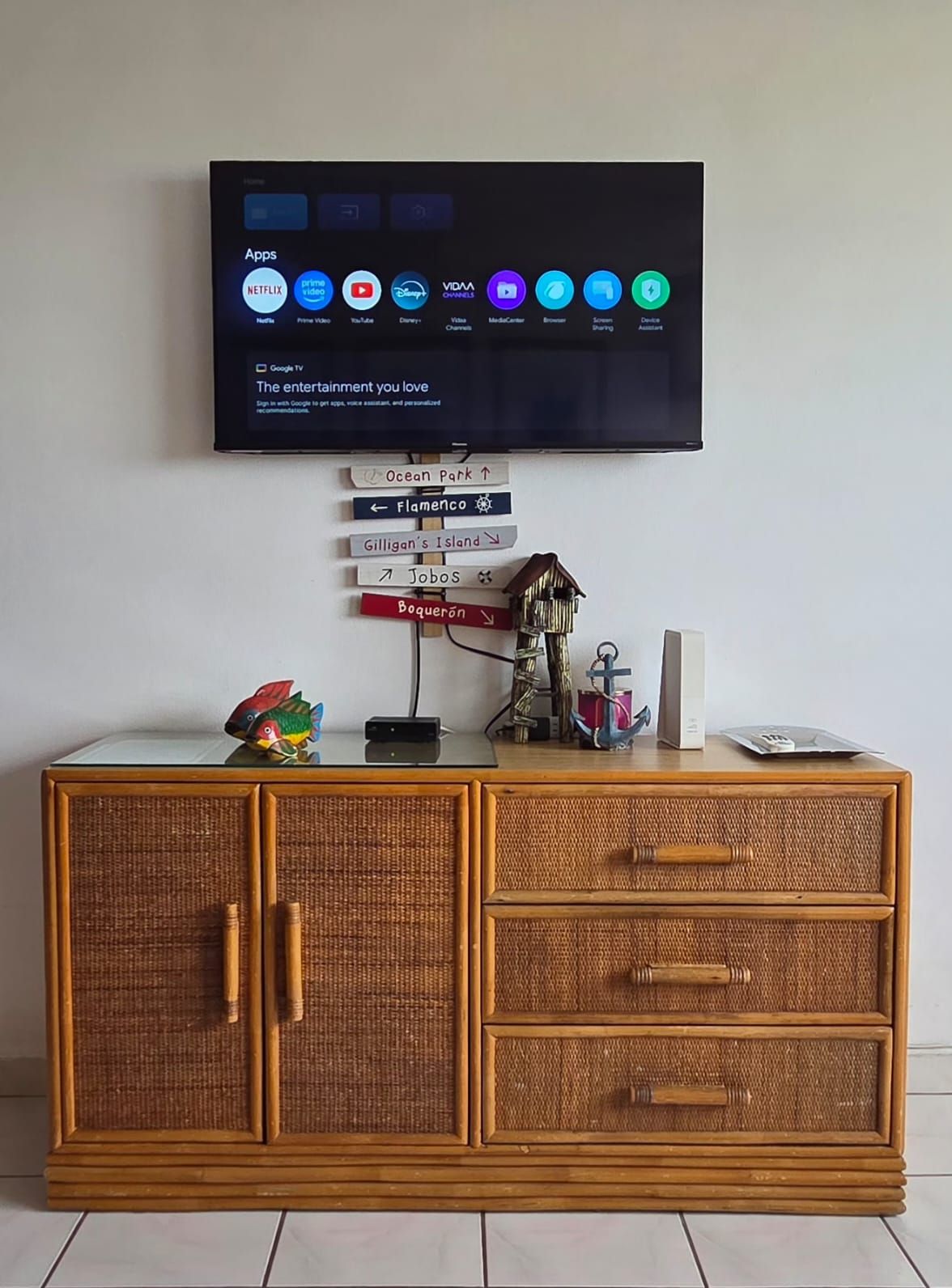 Un televisor montado en la pared muestra varias aplicaciones de transmisión. Debajo del televisor, hay un letrero decorativo con nombres de lugares como Ocean Park y Flamenco. Una casita para pájaros y una figurita de pez están colocadas sobre un mueble de madera con cajones.