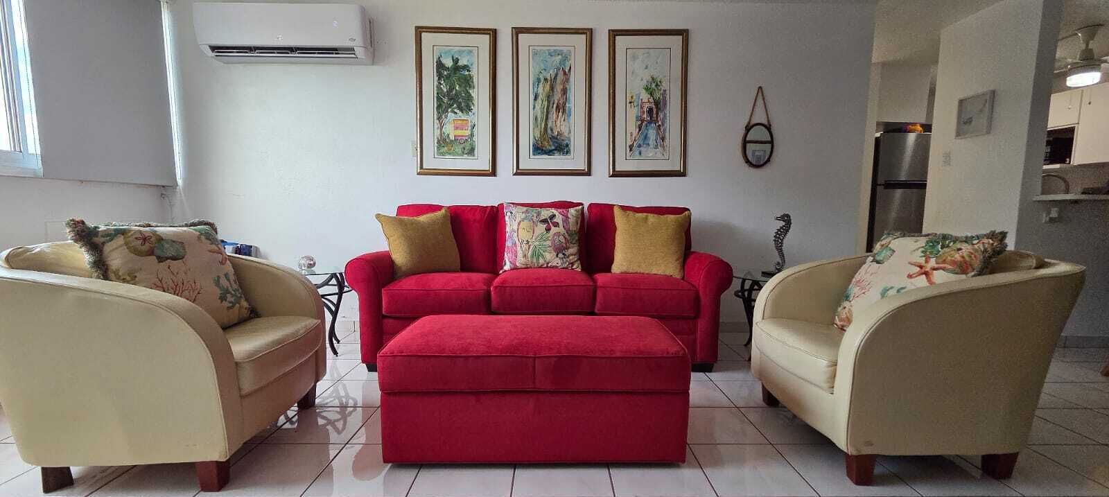 A living room with a red sofa set, beige armchairs, and a matching ottoman. There are three framed artworks on the wall, a hanging mirror, and an air conditioning unit.