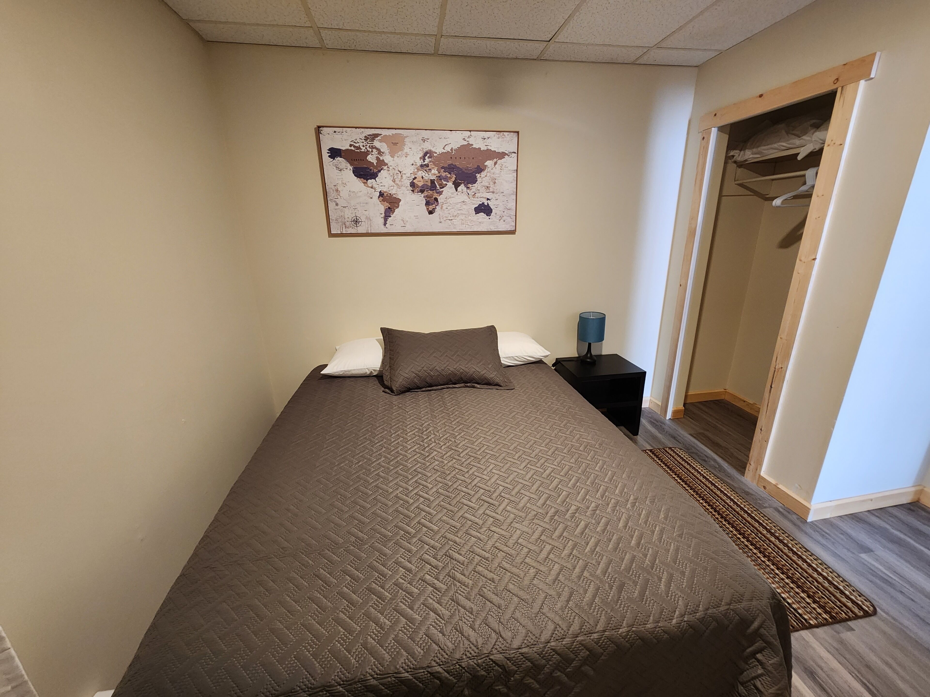 Una cama individual con un edredón marrón, un mapa del mundo en la pared, una mesita de noche con una lámpara y un armario de madera.