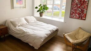Un dormitorio con cama, una mesita de noche de madera, ventana con vista a la calle, planta en maceta y una silla.