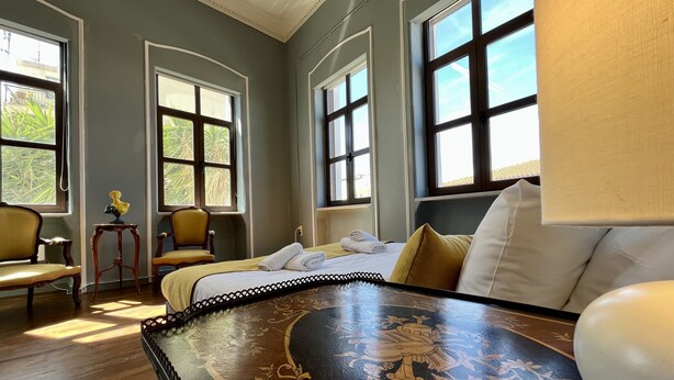 A room with a bed, a table with a decorative design, two chairs, and large windows with views of greenery. - The Thirty8 Heritage House (Chania)