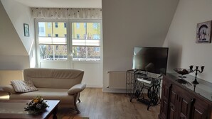 Een woonkamer met een bank, een bijzettafeltje met bloemen, een tv op een dressoir en een kast met een kandelaar.