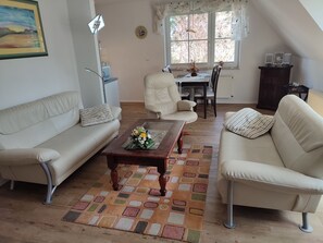 Una sala de estar con sofá, sillón, mesa de centro y mesa de comedor con sillas. Hay una ventana con cortinas, una lámpara y un cuadro en la pared.