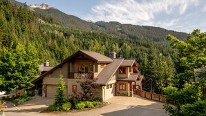 A two-story log cabin with a stone foundation, a wooden deck, and a mountain backdrop. - Ski-In/Ski-Out | Spacious 4BR | Hot Tub | Gear Storage Garage | Managed by Aloha Whistler (Whistler)