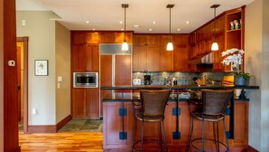 A modern kitchen with wooden cabinets, a stainless steel refrigerator, a microwave, and a bar area with two wicker chairs. - Ski-In/Ski-Out | Spacious 4BR | Hot Tub | Gear Storage Garage | Managed by Aloha Whistler (Whistler)