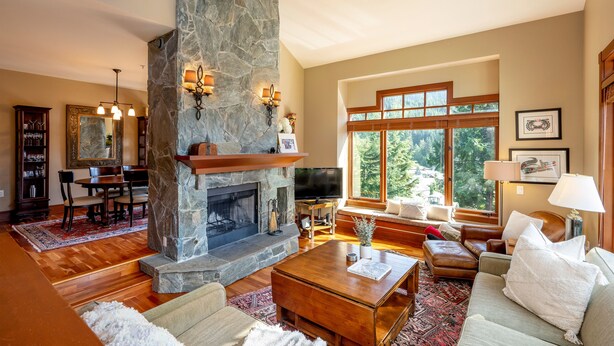 A spacious living room with a stone fireplace, wooden furniture, and large windows offering a view of the outdoors. - Ski-In/Ski-Out | Spacious 4BR | Hot Tub | Gear Storage Garage | Managed by Aloha Whistler (Whistler)